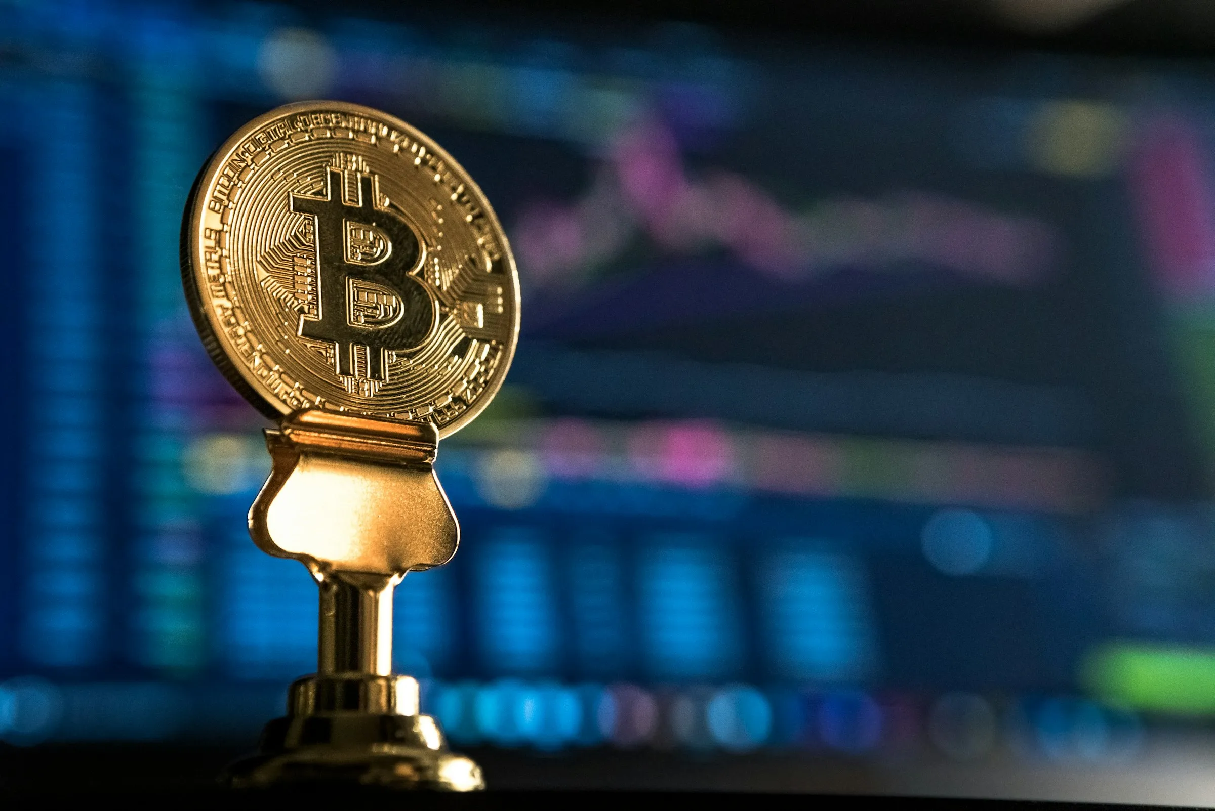 Bitcoin monēta uz statīva pie monitora ar grafikiem